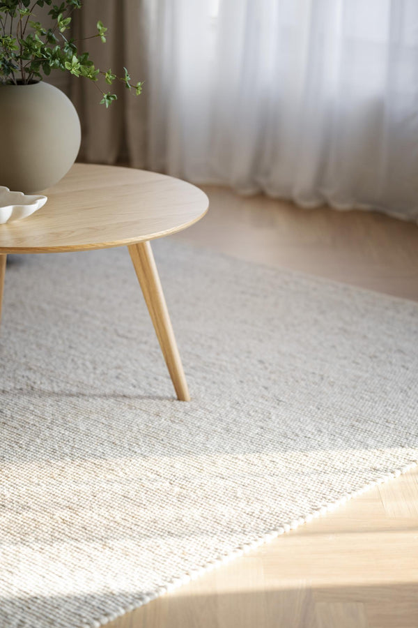 Rund soffbord i ljust trä på vävd matta med beige vas och gröna växter i skandinavisk inredning