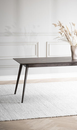 Mörkbrun träbord med sneda ben på vit vävd matta framför vit vägg med tavellist och vas med torkade gräs