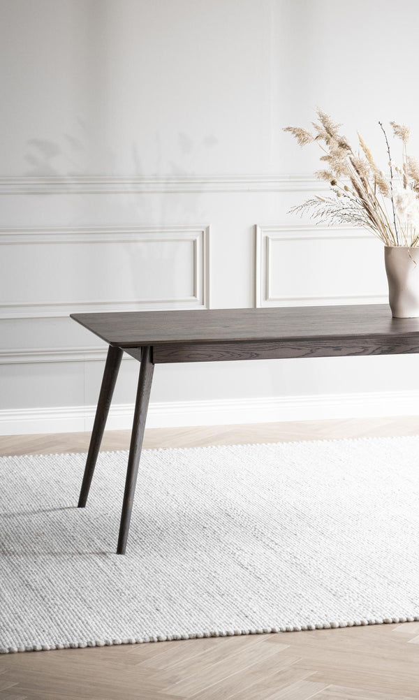 Mörkbrun träbord med sneda ben på vit vävd matta framför vit vägg med tavellist och vas med torkade gräs