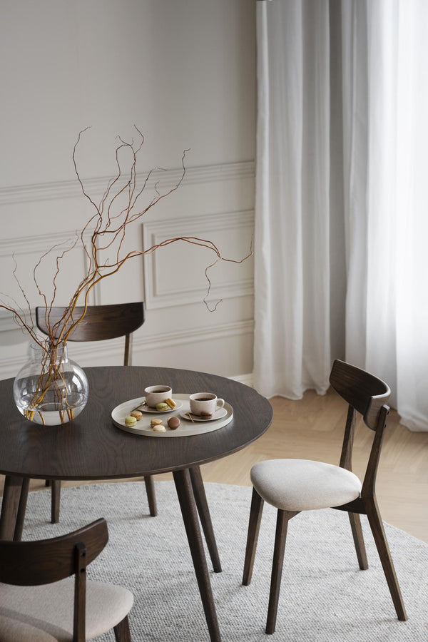 Minimalistiskt matbord i mörk ek med stolar, glasvas med kvistar och kaffeservis i ljust scandinaviskt rum