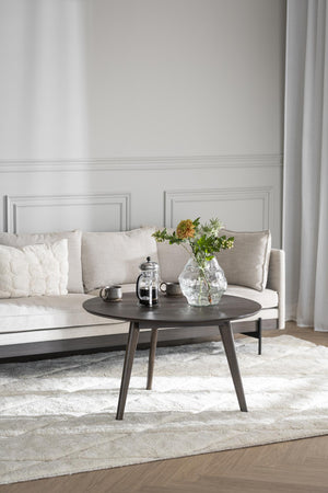 Runt soffbord i mörkt trä med french press och vas med blommor i ljust vardagsrum med beige soffa