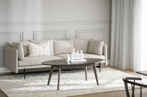 Beige soffa med kuddar och ovalt soffbord i trä på vit matta i ljust vardagsrum med panelvägg