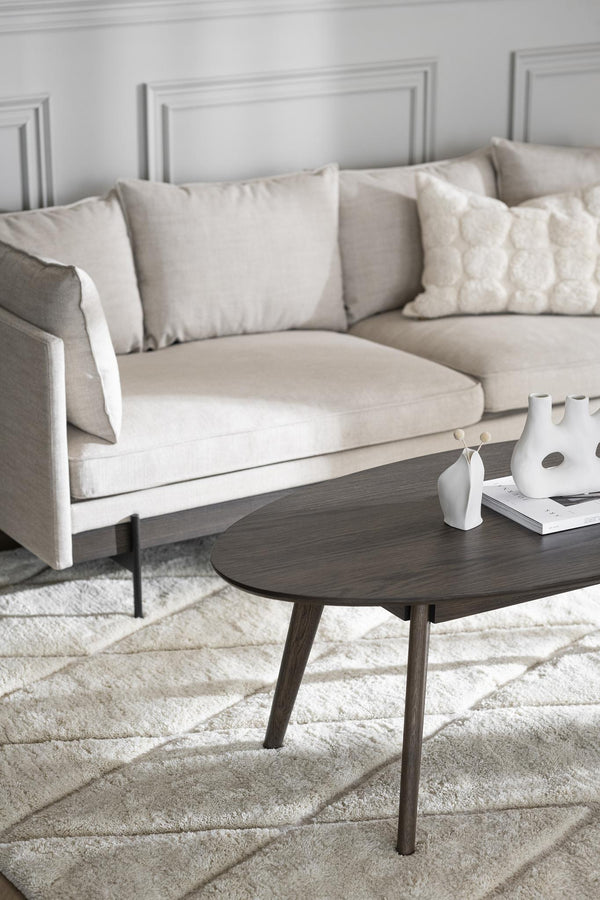 Beige soffa med ljusa kuddar och runt soffbord i mörkt trä på vit matta i vardagsrum