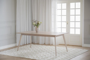Ljust matbord i trä med sneda ben på vit quiltad matta framför fönsterdörr med vita gardiner
