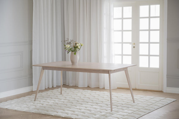 Ljust matbord i trä med sneda ben på vit quiltad matta framför fönsterdörr med vita gardiner