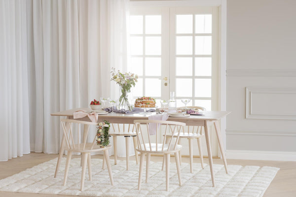 Ljust matrum med träbord, stolar och vackert dukad frukost framför franskt fönster med vita gardiner