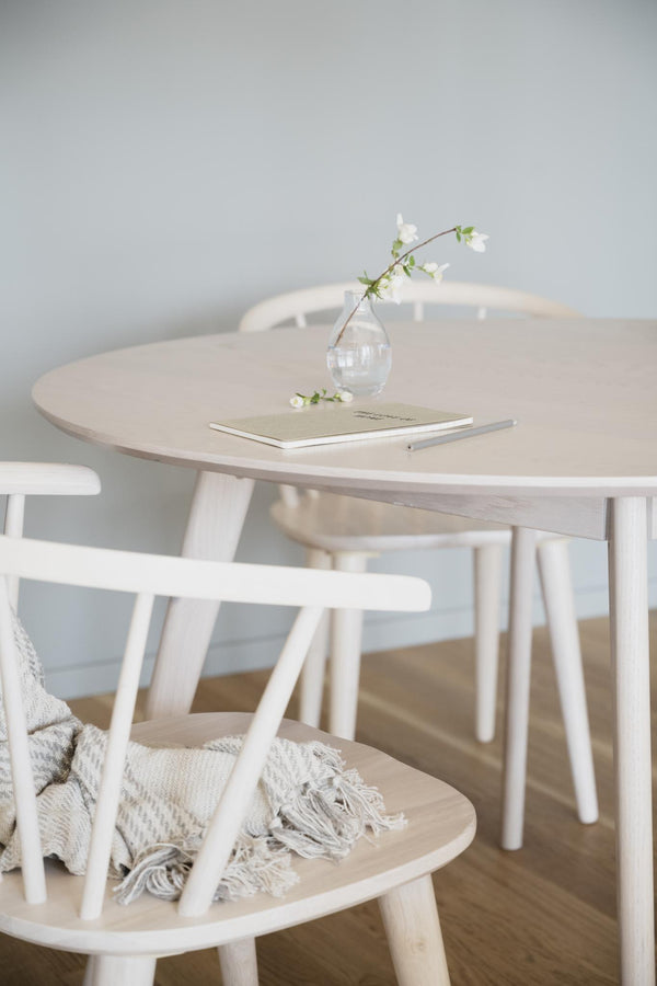 Runt matbord i ljust trä med vita pinnstolar och vas med vita blommor i skandinavisk stil