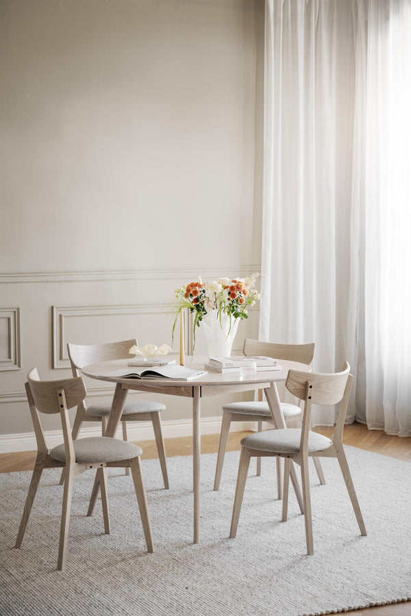 Ljust matbord i trä med fyra stolar och beige klädsel i skandinavisk matsal med vita gardiner