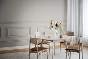 Rund matbord i ljust trä med fyra stolar och bruna dynor i scandinavisk matsalsmiljö med vit vägg