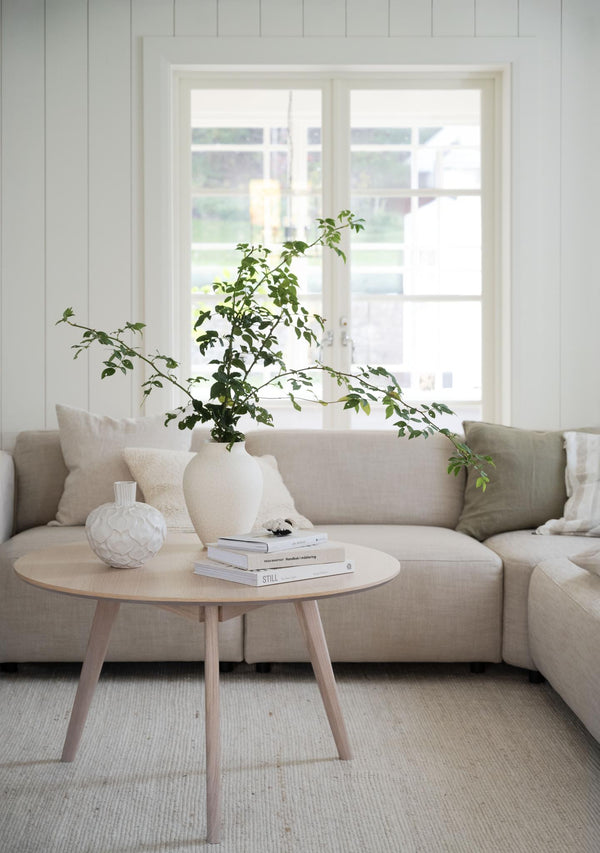 Ljust vardagsrum med beige soffa, runt soffbord i trä och stor vit vas med gröna kvistar framför fönster