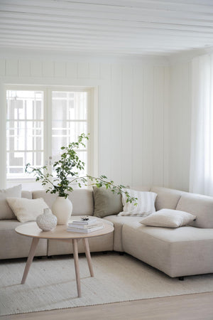 Ljust vardagsrum med beige hörnsoffa, runt soffbord i trä och gröna växter vid fönster