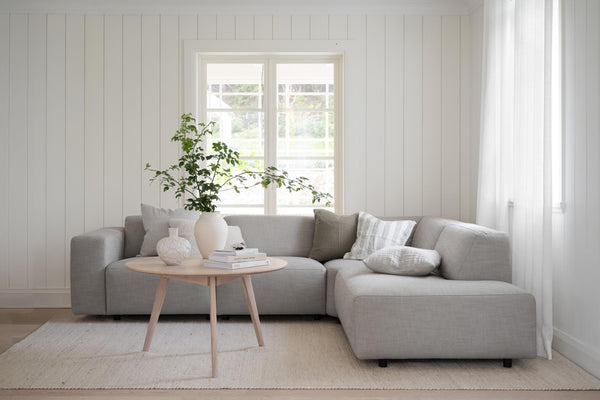 Grå modulsoffa med schäslong i ljust vardagsrum med vit väggpanel och runt soffbord i trä