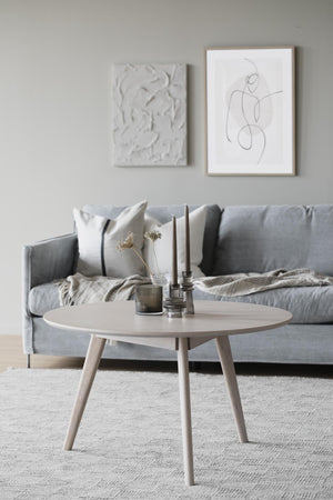 Runt soffbord i ljust trä med tre ben framför grå sammetssoffa i skandinavisk inredningsstil