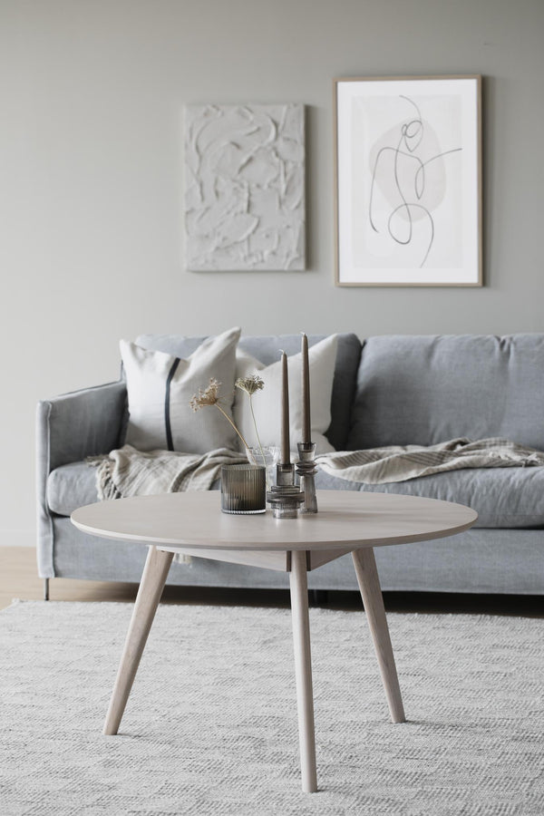 Runt soffbord i ljust trä med tre ben framför grå sammetssoffa i skandinavisk inredningsstil