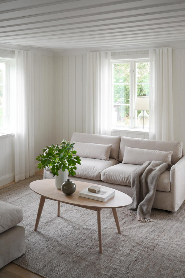 Ljust vardagsrum med beige soffa, ovalt soffbord i trä och vita gardiner framför fönster