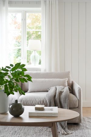 Ljust vardagsrum med beige soffa, rund soffbord i trä, grön växt och fönster med vita gardiner