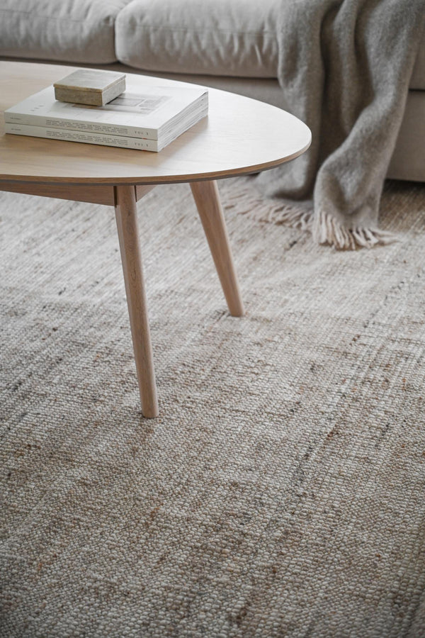 Runt soffbord i ljust trä med böcker på vit luggmatta framför grå soffa med filt