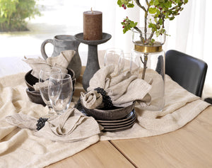 Bordsdukning med beige linneservetter, svarta skålar, vattenglas, ljusstake och grön växt i glasvas