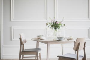 Ljust matsalsbord i trä med stolar, vacker blomstervas och elegant dukning mot vit panelvägg