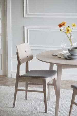 Ljus trämatstol med grå sits bredvid runt matbord i skandinavisk stil med vita väggar i bakgrunden