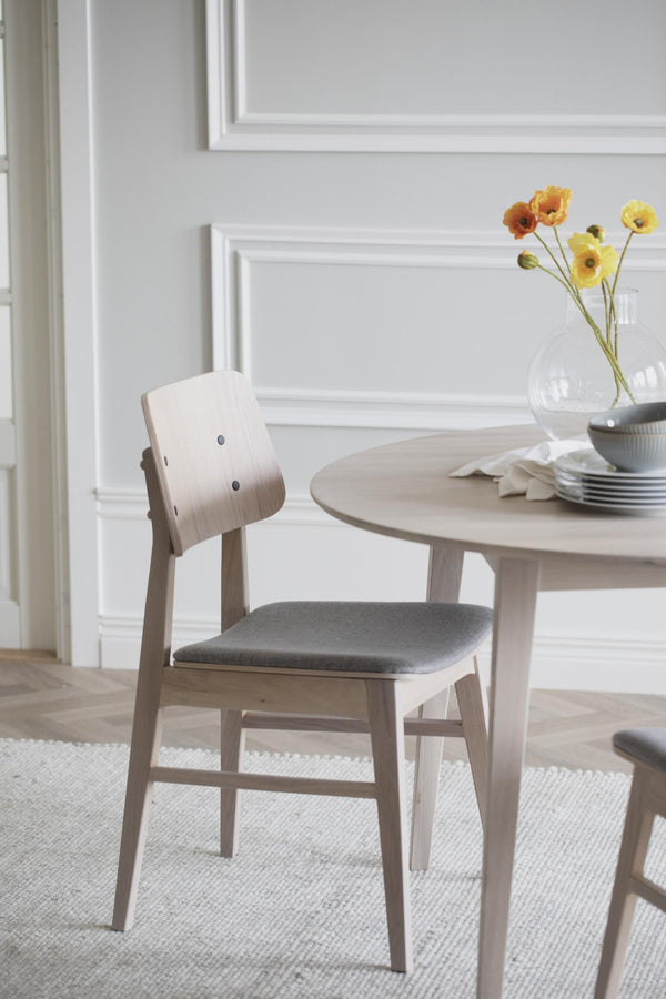 Ljus trämatstol med grå sits bredvid runt matbord i skandinavisk stil med vita väggar i bakgrunden