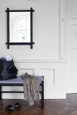 Sittbänk med grå dyna och svart metallram i hall med spegel och vitpanelad vägg