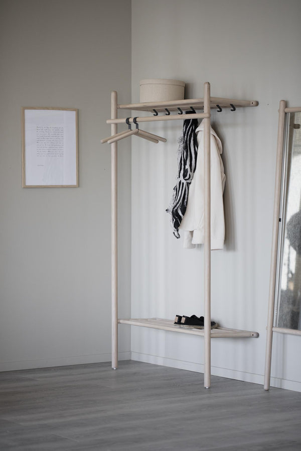 Minimalistisk klädhängare i ljust trä med hylla, krokar och hängande kläder mot vit vägg