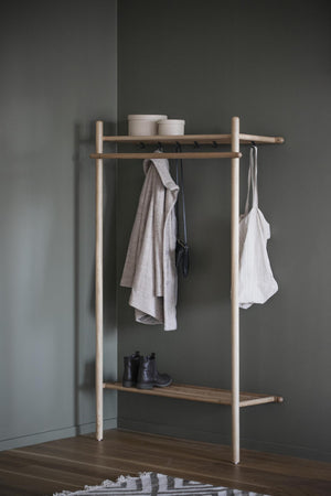 Minimalistisk klädhängare i trä med hyllor, krokar och hängande kläder mot grå vägg