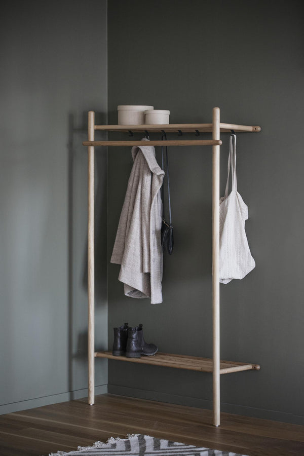 Minimalistisk klädhängare i trä med hyllor, krokar och hängande kläder mot grå vägg