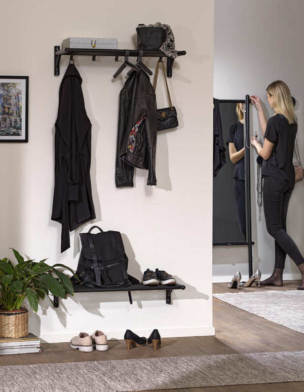 Svart hallmöbel med klädhängare, hylla och spegel där kvinna arrangerar kläder och accessoarer