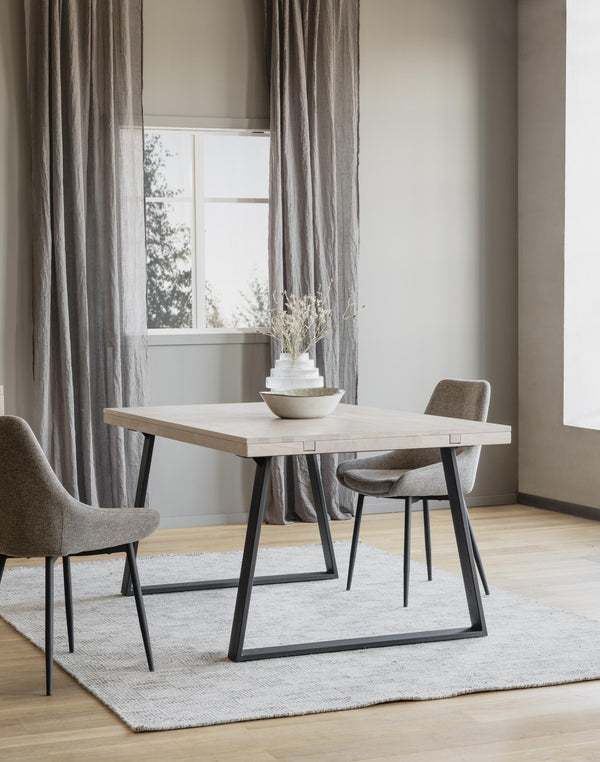 Matbord med ljus träskiva och svarta metallben, två grå stolar, vas med torkade blommor, fönster med gardiner