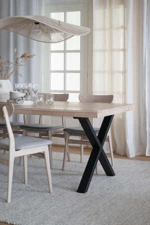 Matbord med ljust trä och svarta krysskors ben, omgivet av stolar i skandinavisk stil med grå dynor