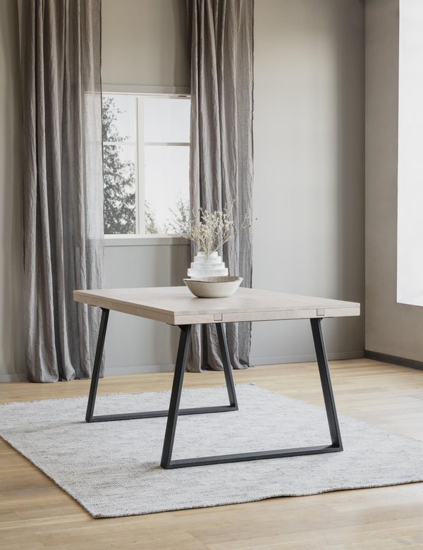 Matbord med ljust trä och svarta metallben på ljus matta framför fönster med grå gardiner