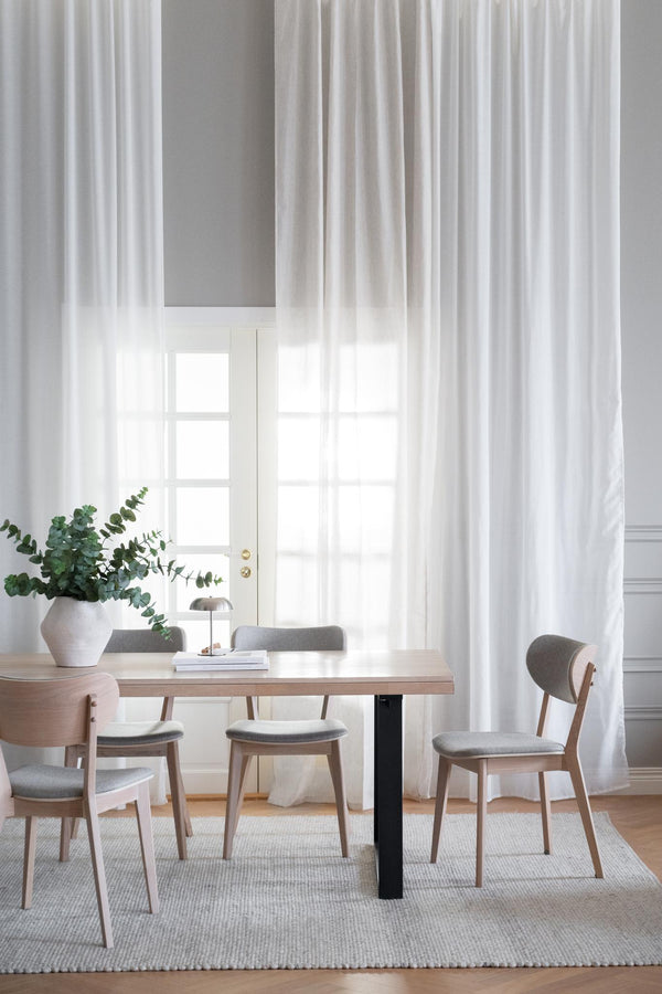 Matbord i ljust trä med gråa stolar och vita gardiner i modern skandinavisk matsal