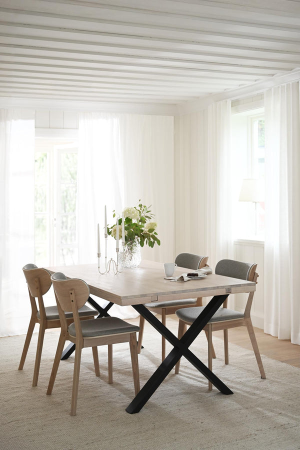 Matbord med krysskena i ljust trä och svart metall, omgivet av stolar med grå stoppad sits i ljust matrum