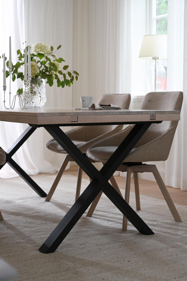 Matbord med X-formade svarta ben och grå skiva, omgivet av beige stolar i modern skandinavisk matsal