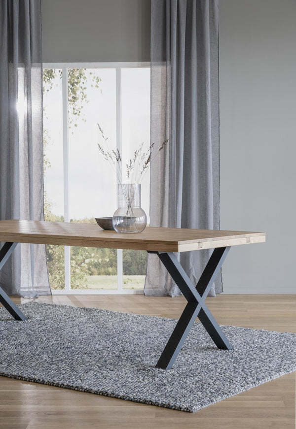 Matbord i ljust trä med svarta kryss-ben på grå matta framför fönster med gardiner