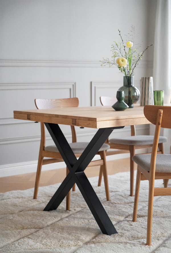 Matbord med ljus ekskiva och svart X-formad metallstomme, omgivet av trässtolar med grå dynor