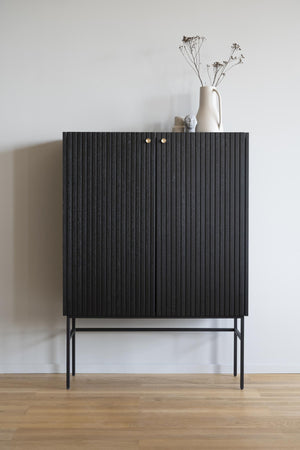 Svart skåp med räfflad yta på smala metallben, dekorerat med vit vas och torkade blommor