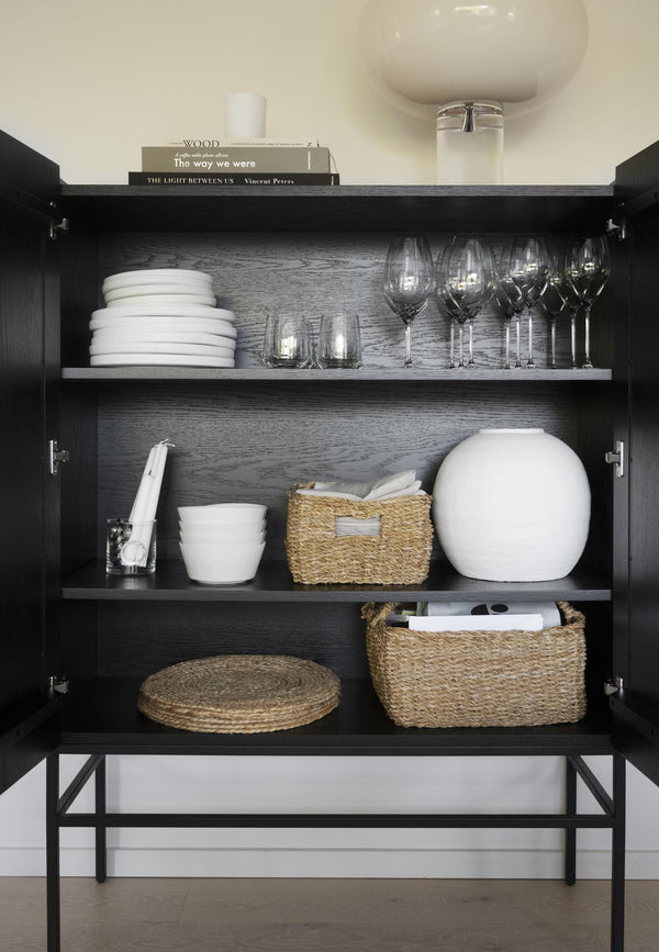 Öppen svart skänk med vita tallrikar, vinglas, rotingkorgar och servering organiserat på hyllor