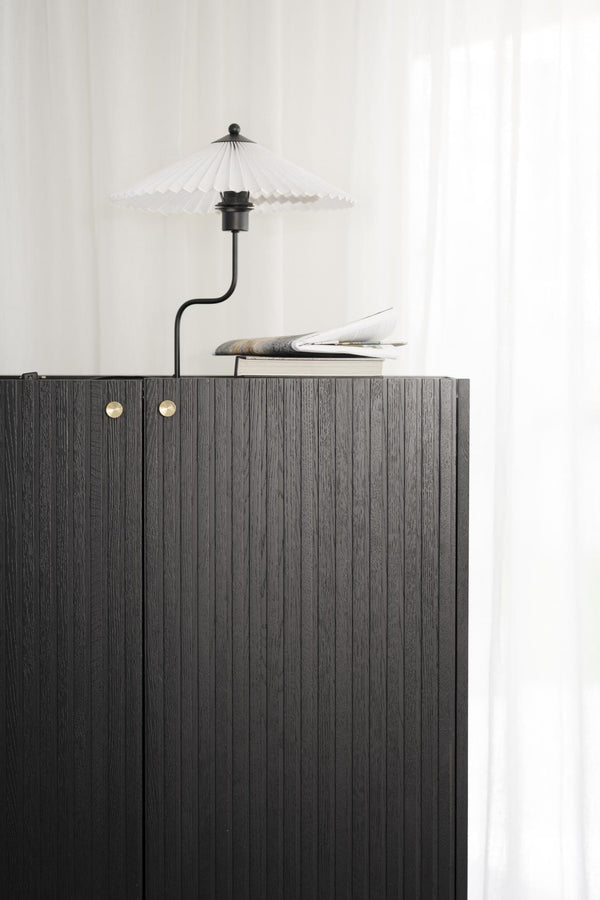 Svart skänk med räfflad yta och mässingsdetaljer, dekorerad med lampa och böcker mot vit vägg