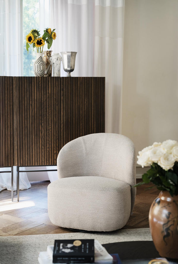 Beige fåtölj i teddy-tyg framför mörkt träskåp med dekorativa vaser och blommor i elegant vardagsrum