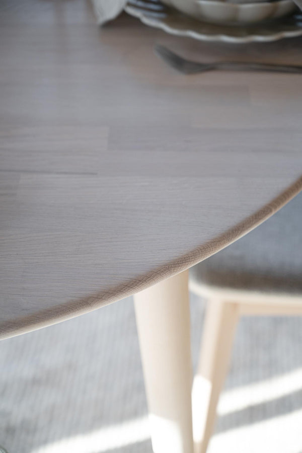 Närbild på ljust träbord med synlig kant och bordsben i naturligt trä, porslin i bakgrunden