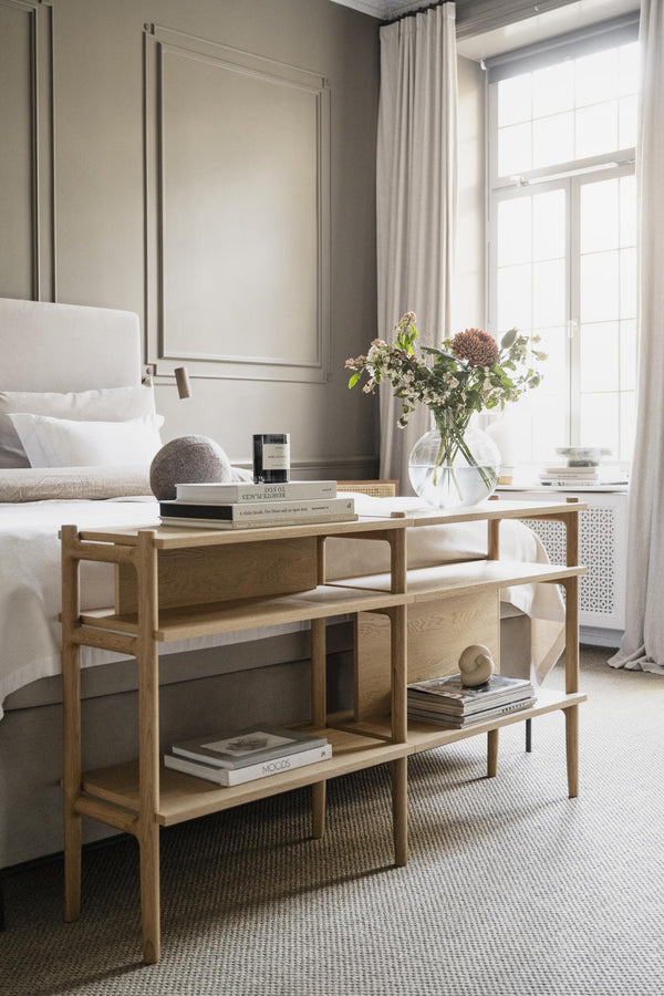 Ljust sovrum med sängbord i ljust trä, dekorerat med böcker, vas med blommor och vit sängkläder