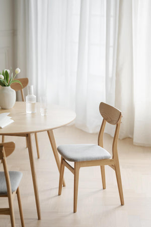 Ljus matsalsmiljö med rund träbord och stolar i skandinavisk design med vita gardiner i bakgrunden