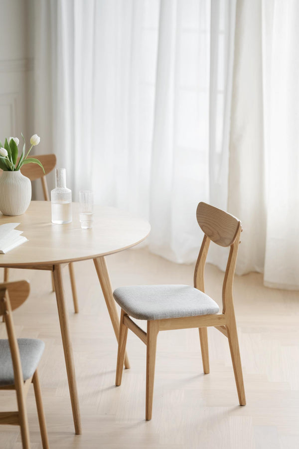 Ljus matsalsmiljö med rund träbord och stolar i skandinavisk design med vita gardiner i bakgrunden