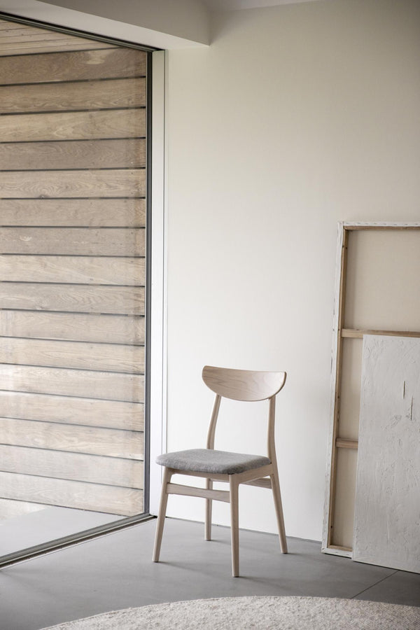 Minimalistisk stol i ljust trä med grå stoppad sits placerad framför stort fönster i skandinavisk interiör