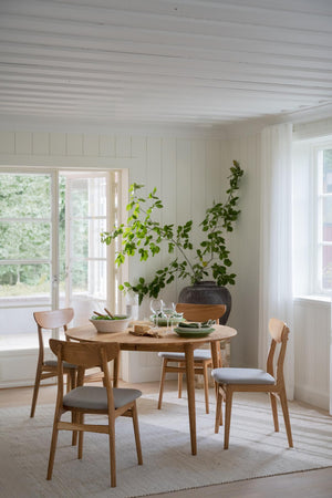 Runt matbord i trä med fyra stolar och grå dynor i ljust skandinaviskt kök med vita väggar