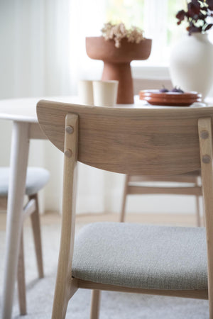 Matstol i ek med grå stoppad sits och böjd rygg, vid matbord med dekorativa föremål