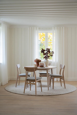 Rund matbord i ljust trä med fyra stolar och grå dynor på ljus rund matta i matsal med fönster